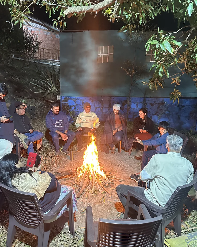 Campers in a circle of chairs around a bonfire on the farm at night, near Rajgad Fort.