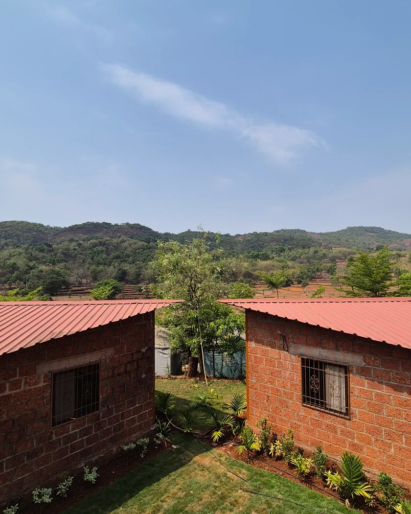 The cottages at Sujalam Sufalam farmstay near Pune, with green Sahyadri hills and a clear sky behind them.