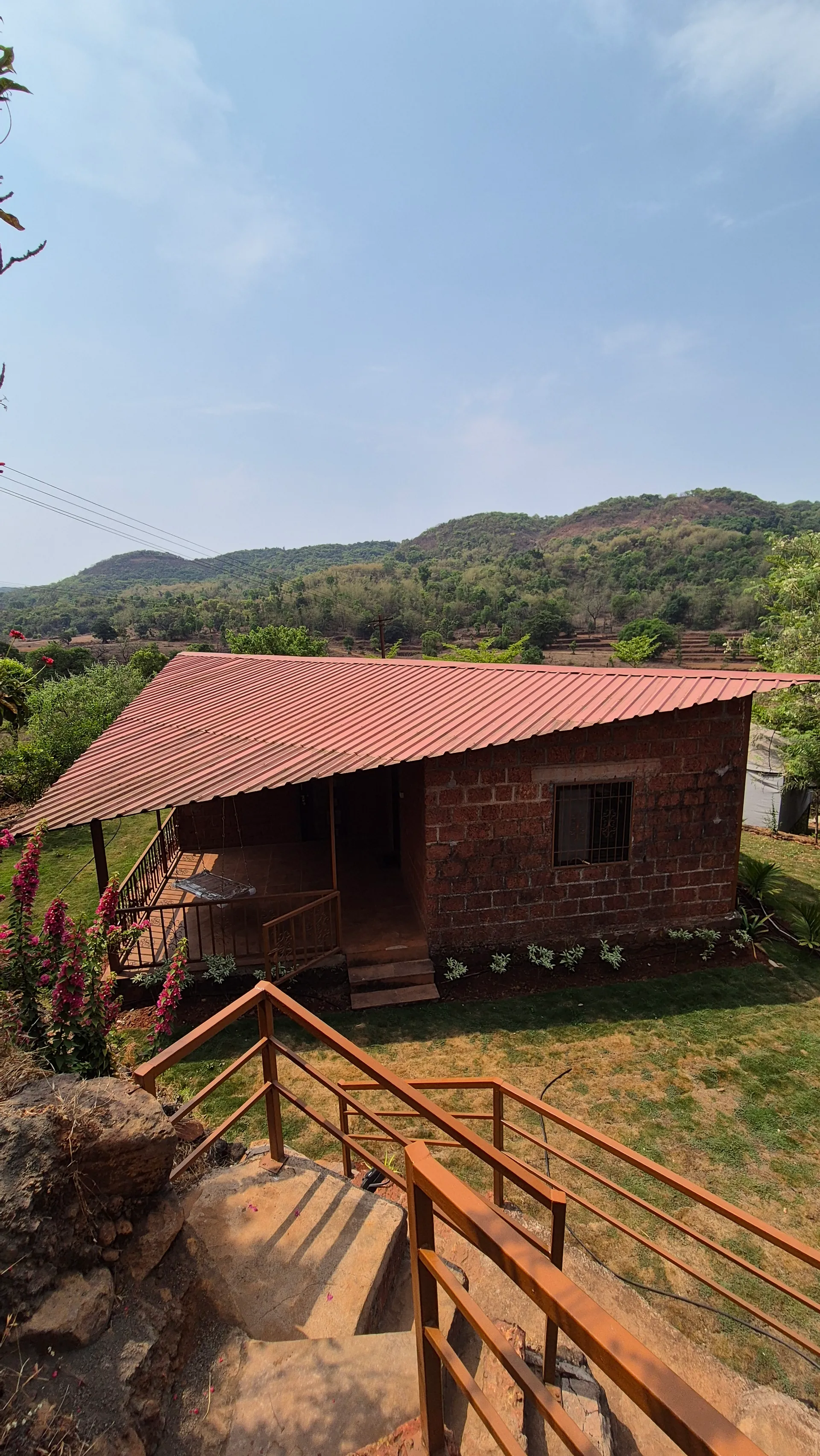 A red-roofed stone cottage on the farm, its verandah facing the lawn, with wooded Sahyadri hills behind.