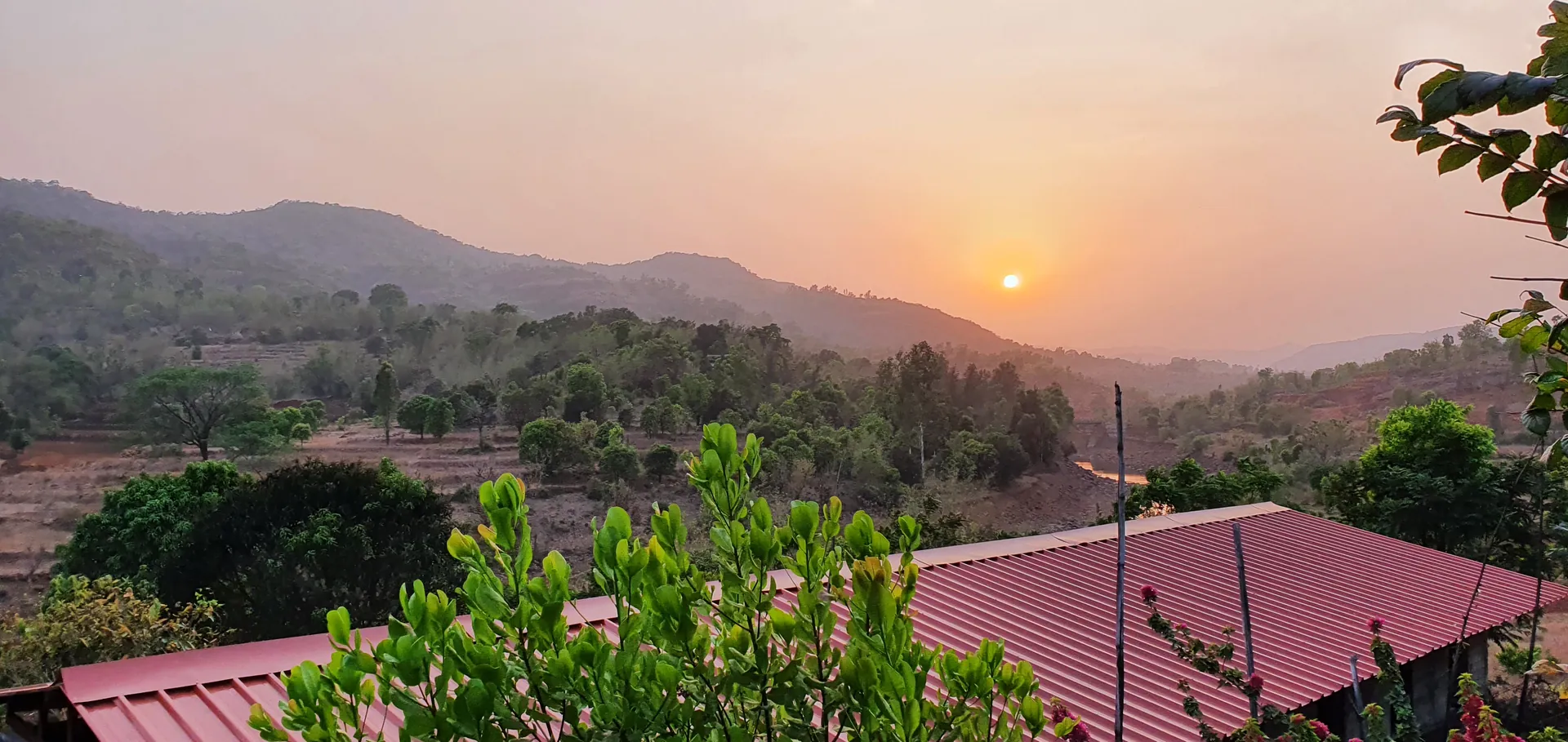 The sun going down over the hills, seen past the red roof of a cottage.